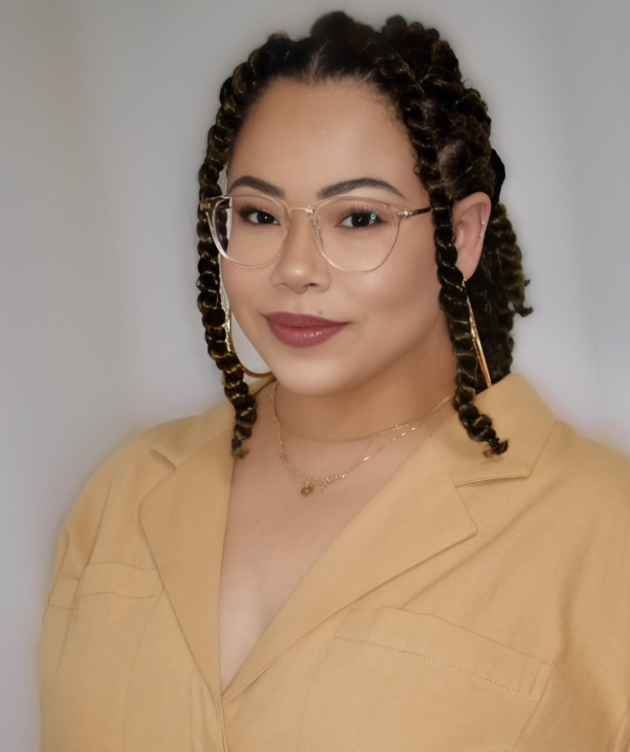 A photo of a woman with light skin and dark hair twists in clear-framed glasses, a beige collared top and gold necklaces