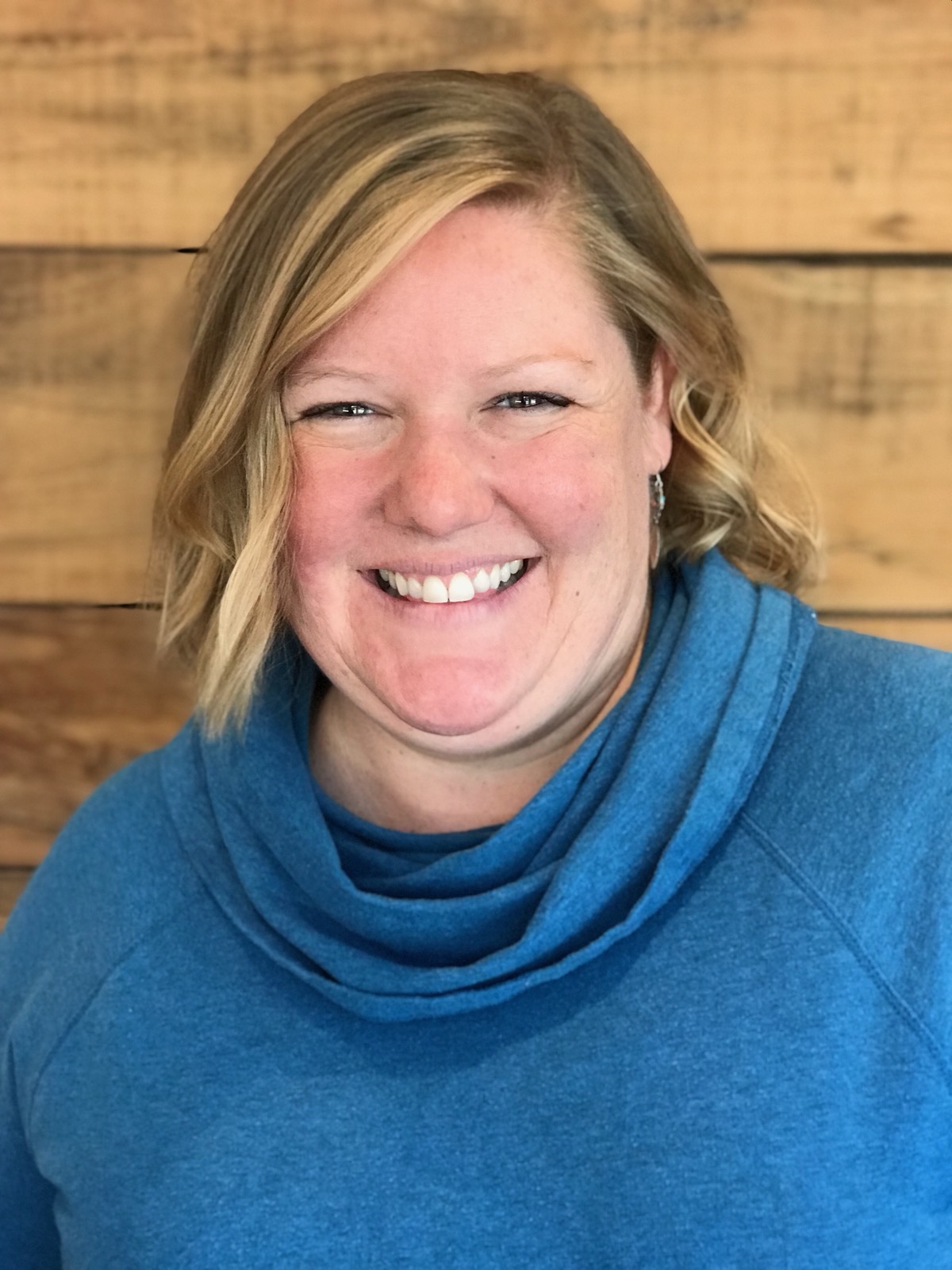 Woman with blonde hair standing in front of a wooden wall smiling in a blue top