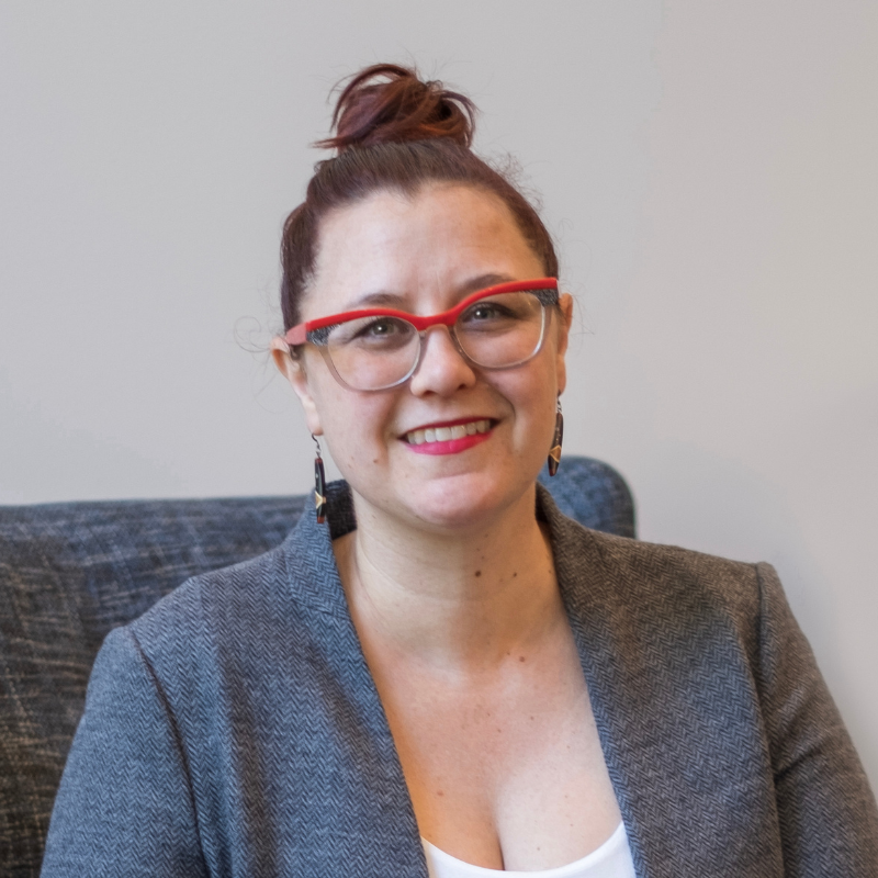 The photo shows Erin Allgood, a white woman with dark hair pulled back into a bun. She is wearing glasses with bright red frames, a blazer, and a white shirt. 