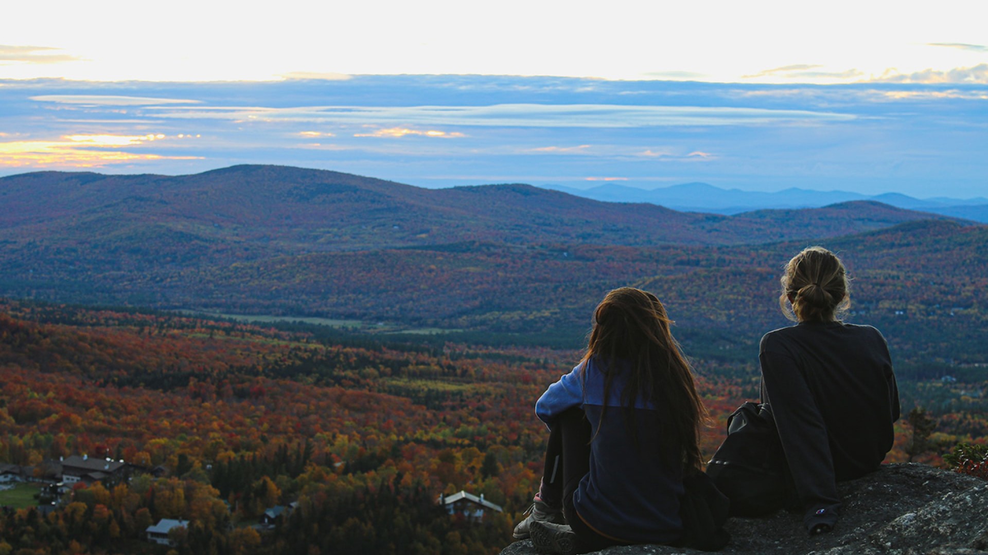 Two individuals sitting on a rock looking out over fall foliage mountain view. You can see the backs of the people